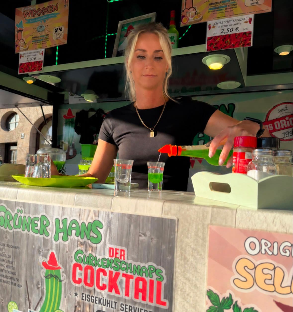 Angeklebtes Foto: Ausschank am Gurkenschnaps-Wagen „Grüner Hans“ – Gurkenschnaps wird aus der Flasche in Shotgläser eingeschenkt, daneben die Karte mit dem Gurkenschnaps-Cocktail