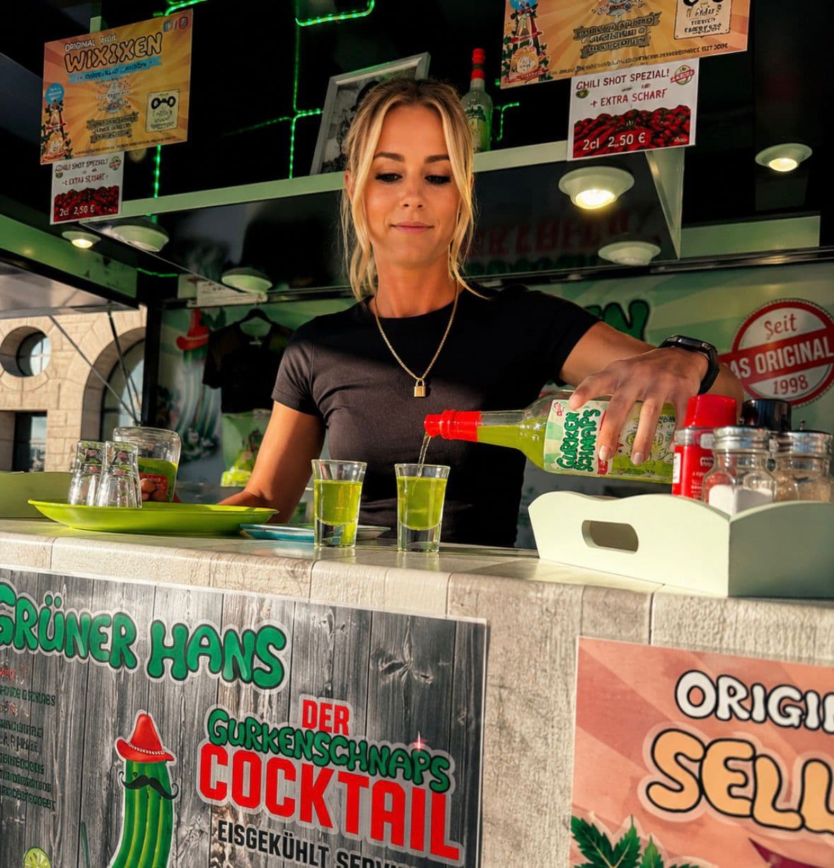 Angeklebtes Foto: Ausschank am Gurkenschnaps-Stand – Gurkenschnaps wird in Shotgläser eingeschenkt